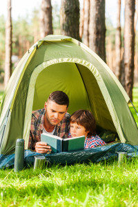 Spending good time outdoors. Father and son reading book while lying in tent together